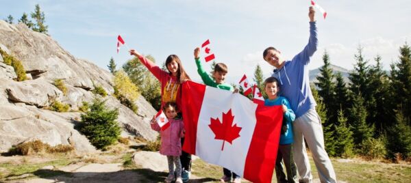 Happy family holding a large Canadian flag outdoors, surrounded by scenic nature, celebrating their move to Canada.