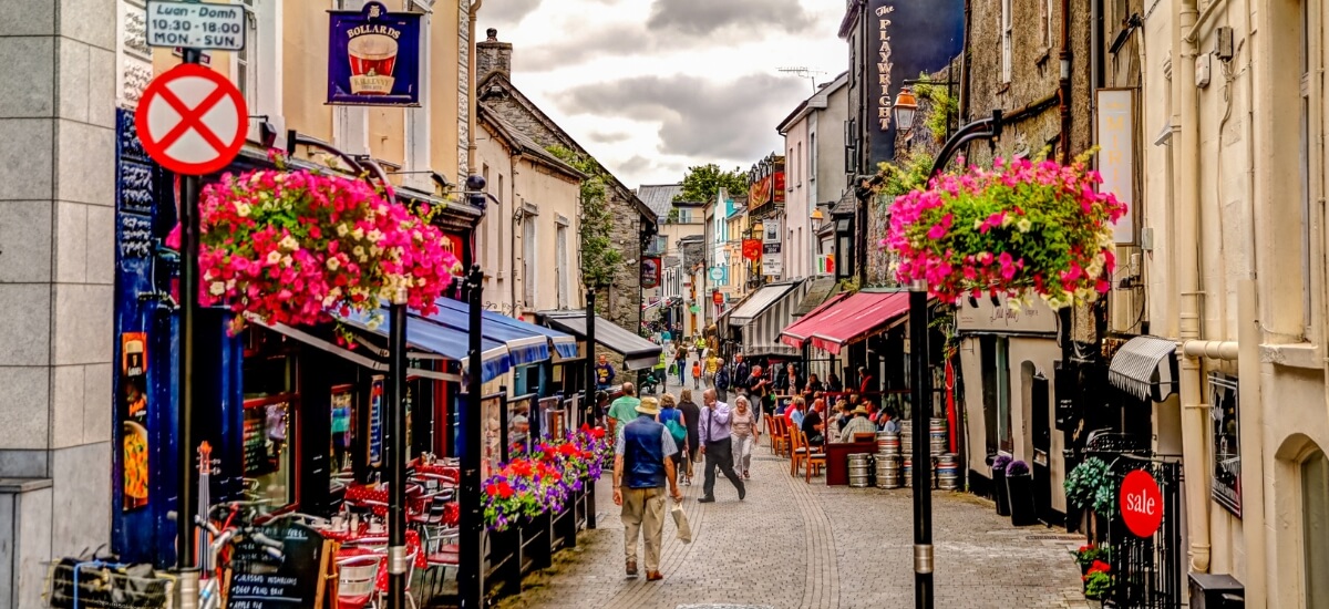 Busy Irish shopping street with colourful storefronts, outdoor dining, and hanging flower baskets, showcasing the lively atmosphere of a typical Irish town. Busy Irish shopping street with colourful storefronts, outdoor dining, and hanging flower baskets, showcasing the lively atmosphere of a typical Irish town.