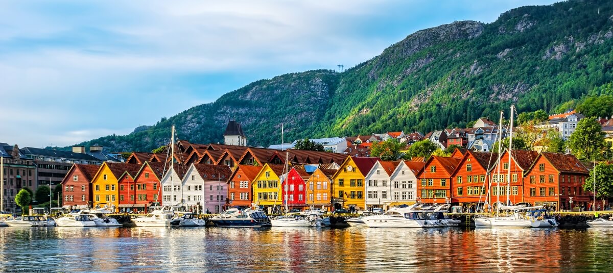 Colourful waterfront buildings in Bergen, Norway, with boats docked along the shore and lush green mountains in the background. Colourful waterfront buildings in Bergen, Norway, with boats docked along the shore and lush green mountains in the background.