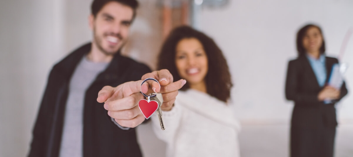 Happy couple holding new home keys in Dublin with heart keyring and estate agent in the background.