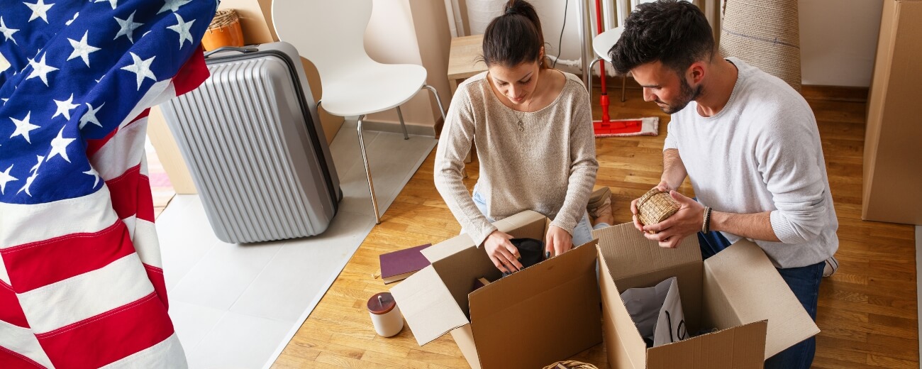 Couple packing boxes together with an American flag in the background, preparing for their move from England to the USA