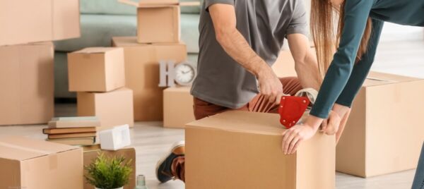 Couple sealing a moving box while preparing for a house move, surrounded by cardboard packing boxes.