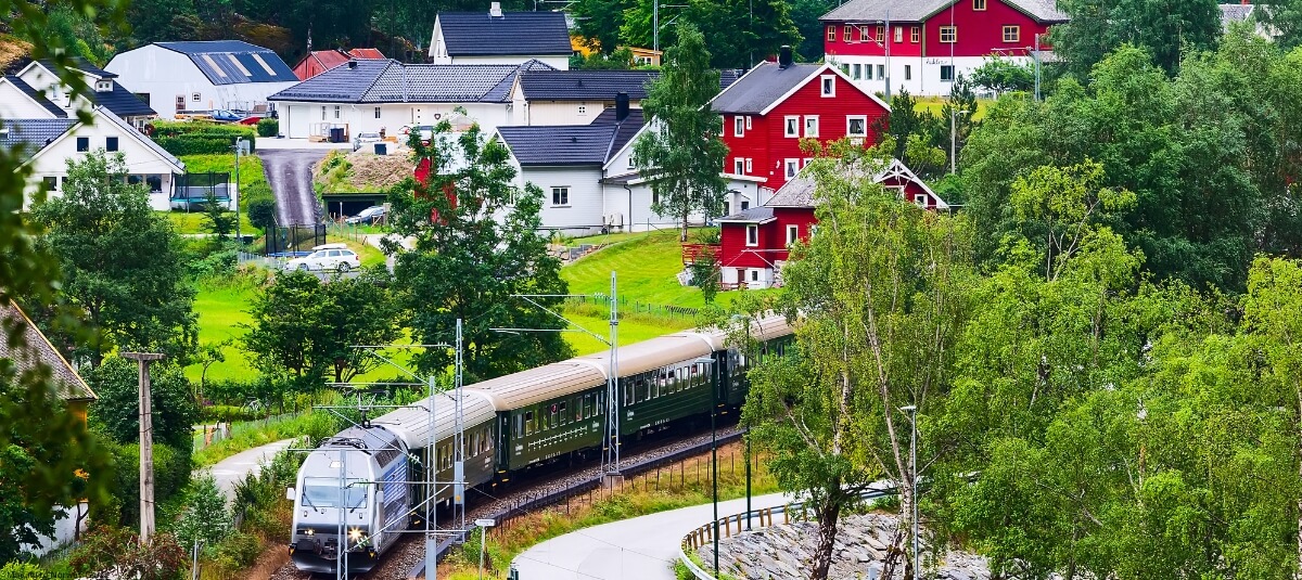 Scenic view of a train passing through a small Norwegian village with red and white houses, surrounded by green trees and hills. Scenic view of a train passing through a small Norwegian village with red and white houses, surrounded by green trees and hills.